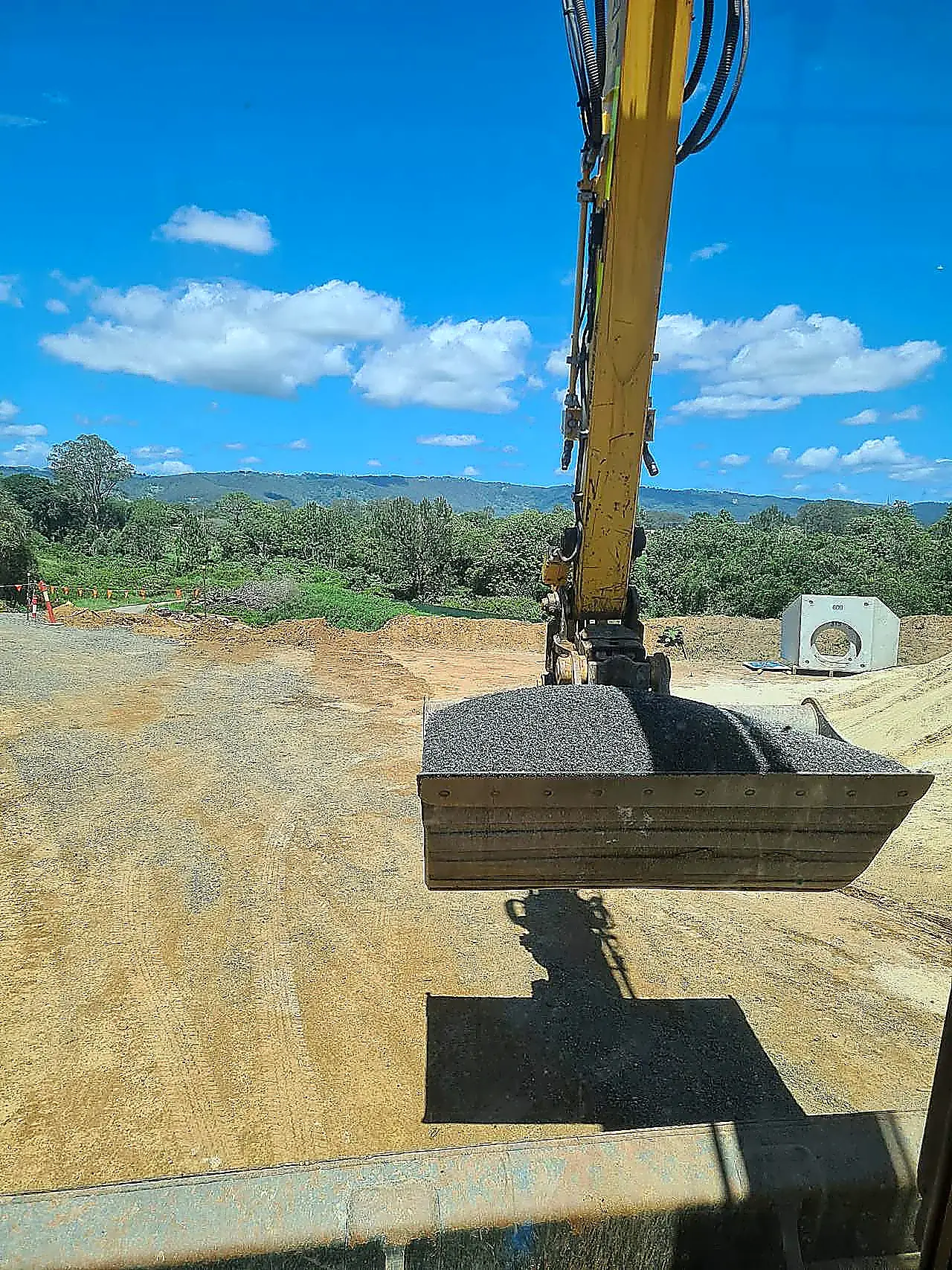 Pipeline bedding gravel and precast pit, SEQ Yellow excavator carrying a bucket of crushed aggregate with a precast concrete pit on an SEQ civil site, photographed from the cab.