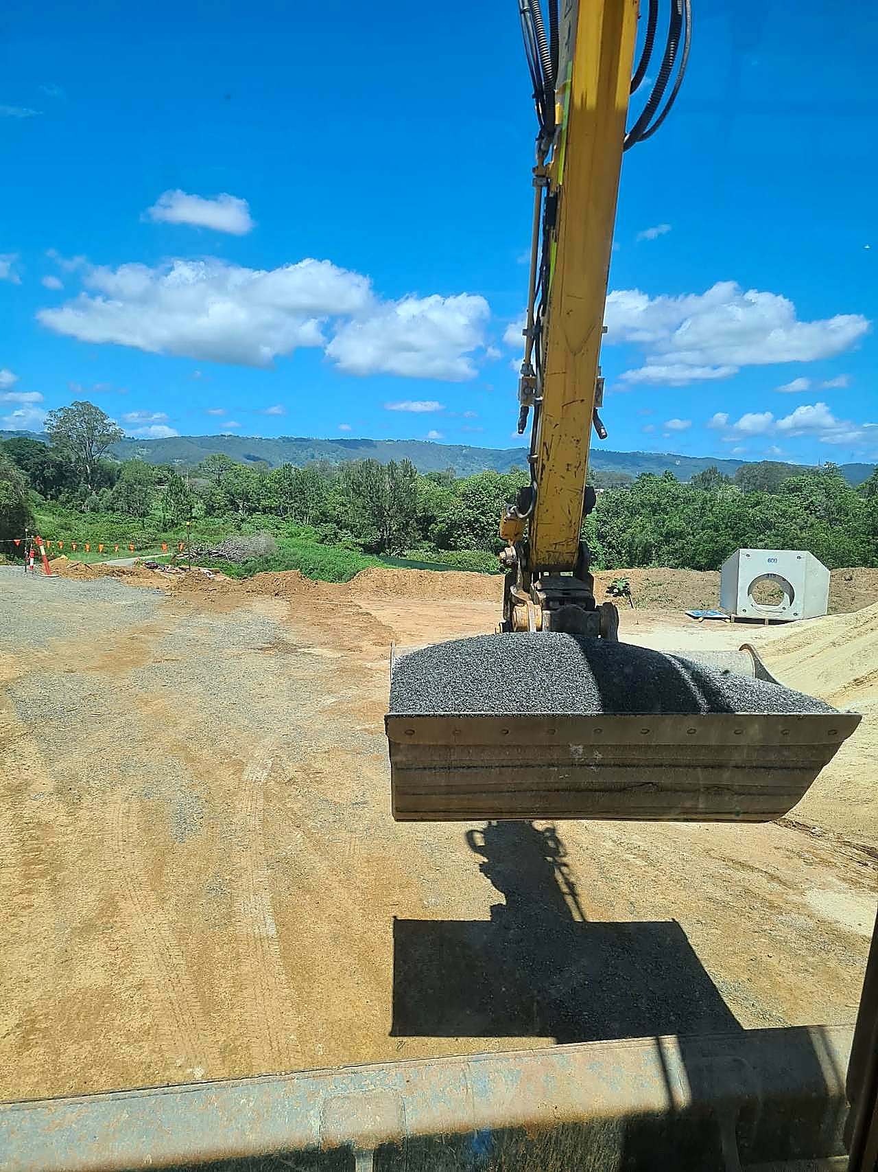 Pipeline bedding gravel and precast pit, SEQ Yellow excavator carrying a bucket of crushed aggregate with a precast concrete pit on an SEQ civil site, photographed from the cab.